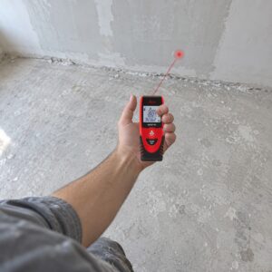 Leica Disto D1 w dłoni podczas pomiaru, kropka lasera na ścianie